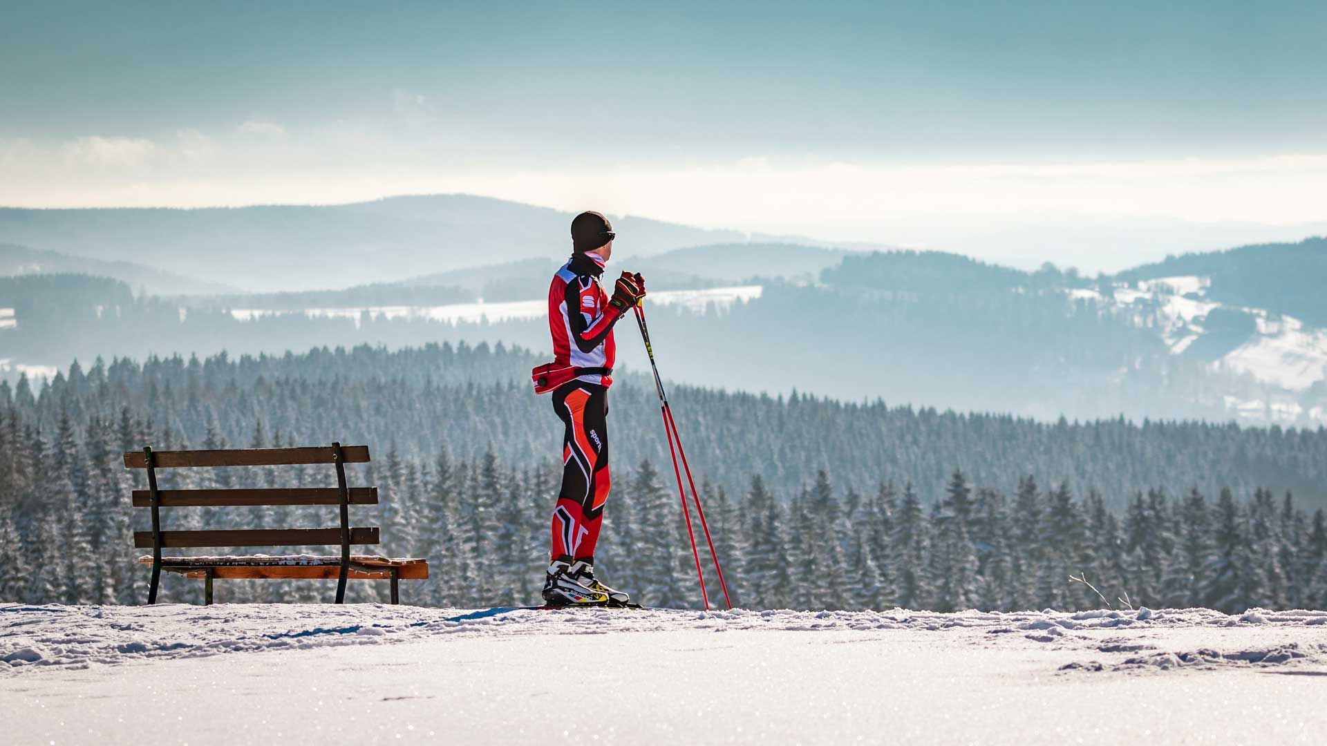 Skifahrer blickt auf verschneiten Wald, neben Bank